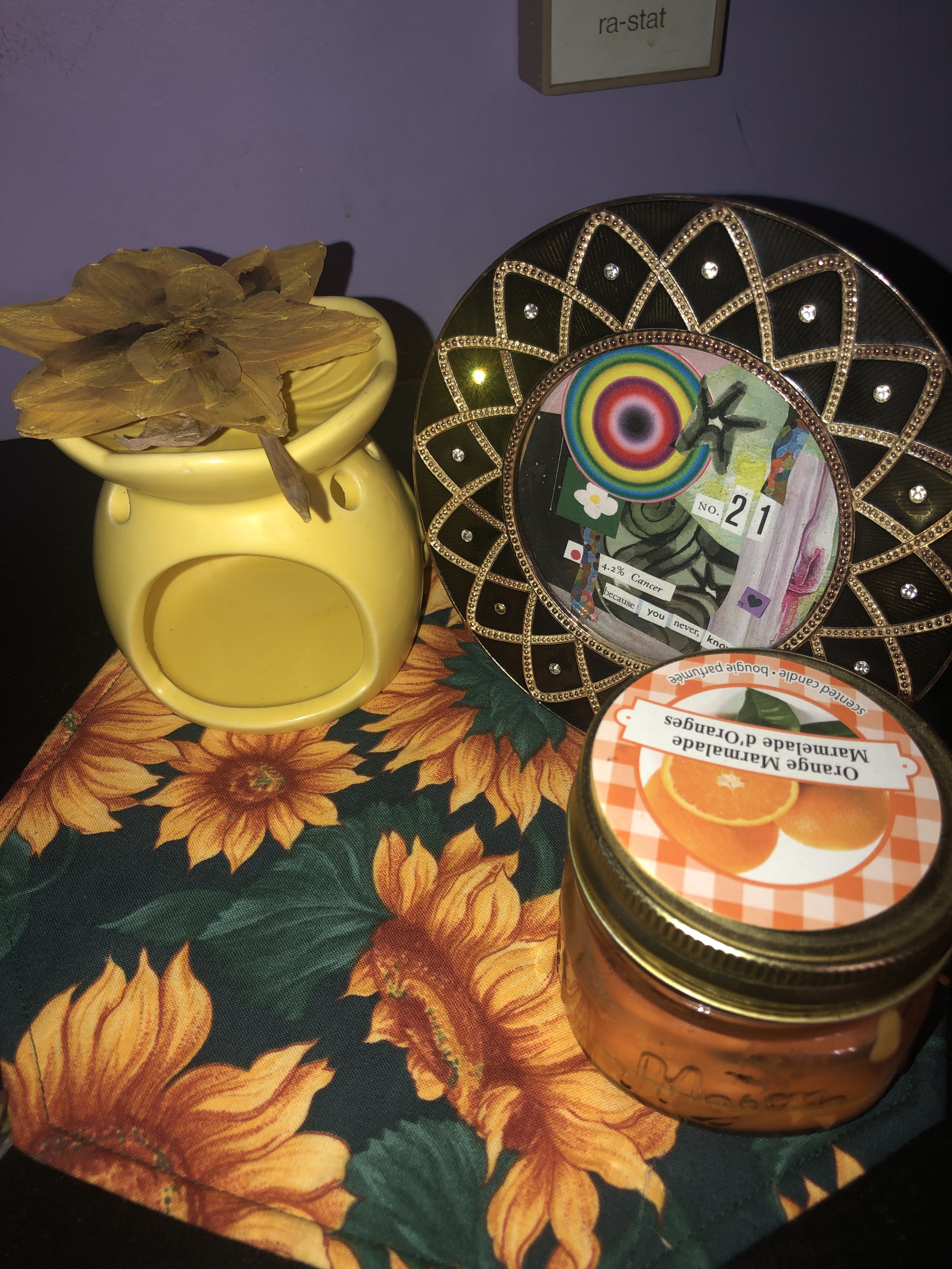 A photo of my altar for Apollo. There is a sunflower placemat. On top of it there is a yellow oil warmer with a pressed daffodil on top. Beside it is a black and gold round picture frame holding a personal collage I've made. In front of the frame is an orange candle in a jar.