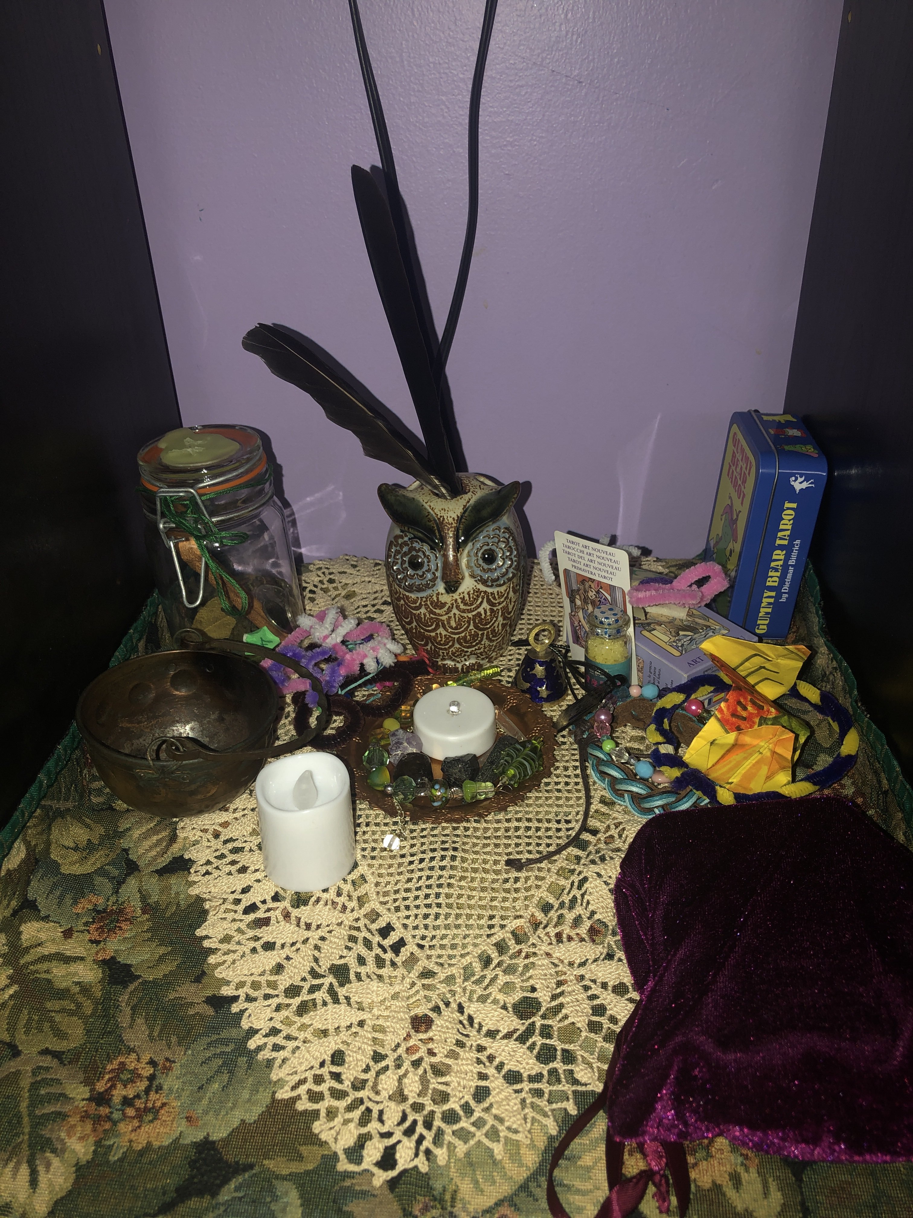 A photo of my altar devoted to Athena. There is a green leaf placemat with a beige lace placemat above it. On the altar there are tarot decks, battery candles, a bowl, jars, an owl figurine with real feathers in it, and various craft objects.
