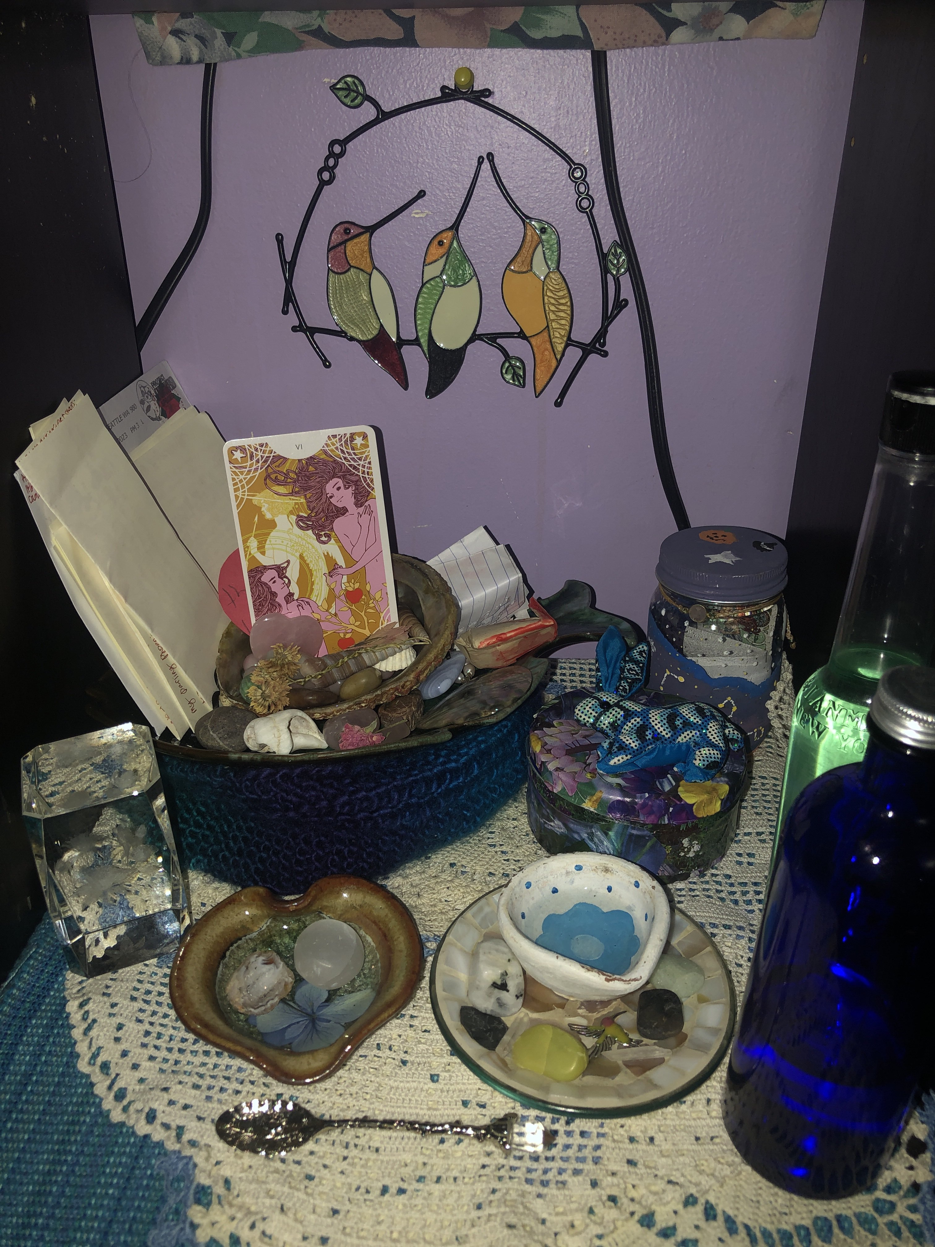 A photo of my altar devoted to Aphrodite. It has a blue placemat alongside white placemats above it. There are various objects on this altar such as a water bowl, various bottles, jars, spools and various crystals. On the wall behind it is a stained glass piece of hummingbirds.